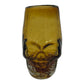 handblown Mexican shot glass, shaped like a skull and crafted from eco-friendly recycled glass. It features a gradient color scheme transitioning from dark at the base to a lighter amber at the top. The glass holds approximately 2 fluid ounces.