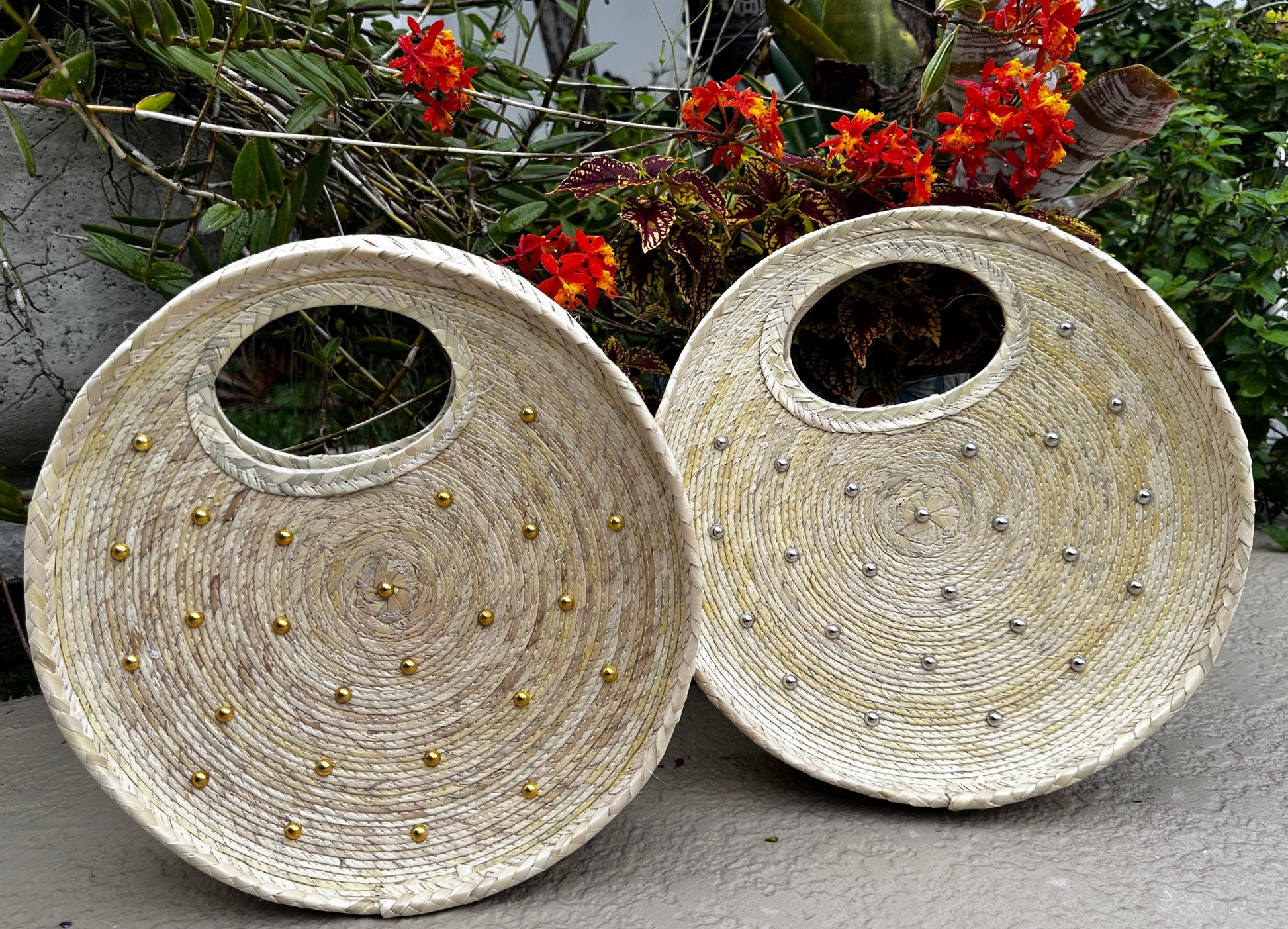 two round, handwoven handbags made of natural fibers. Each bag features a circular handle cut-out and is adorned with metallic studs—one with gold studs and the other with silver studs.