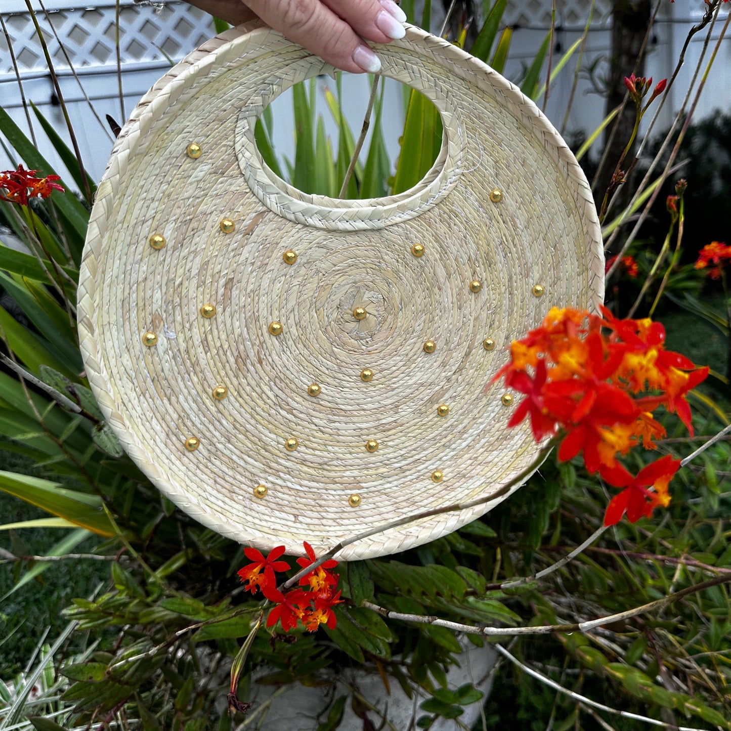 handwoven handbag made of natural fibers. It has a circular handle cut-out at the top and is decorated with small gold metallic studs arranged in a spiral pattern.