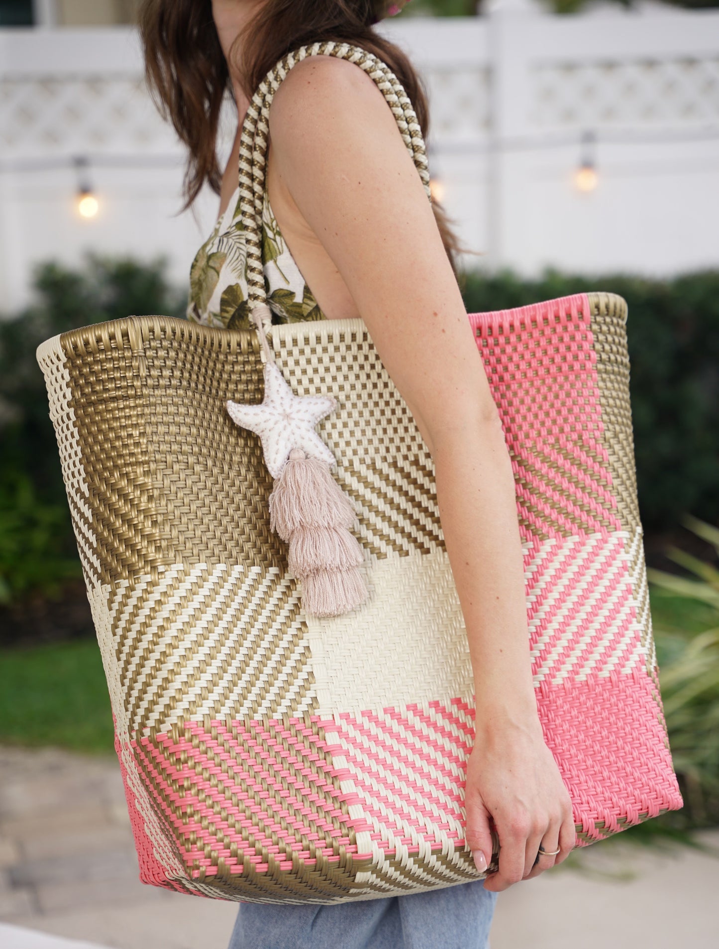 A woman carrying a large woven tote bag with a patchwork design in olive green, cream, and pink, featuring a white starfish tassel charm.