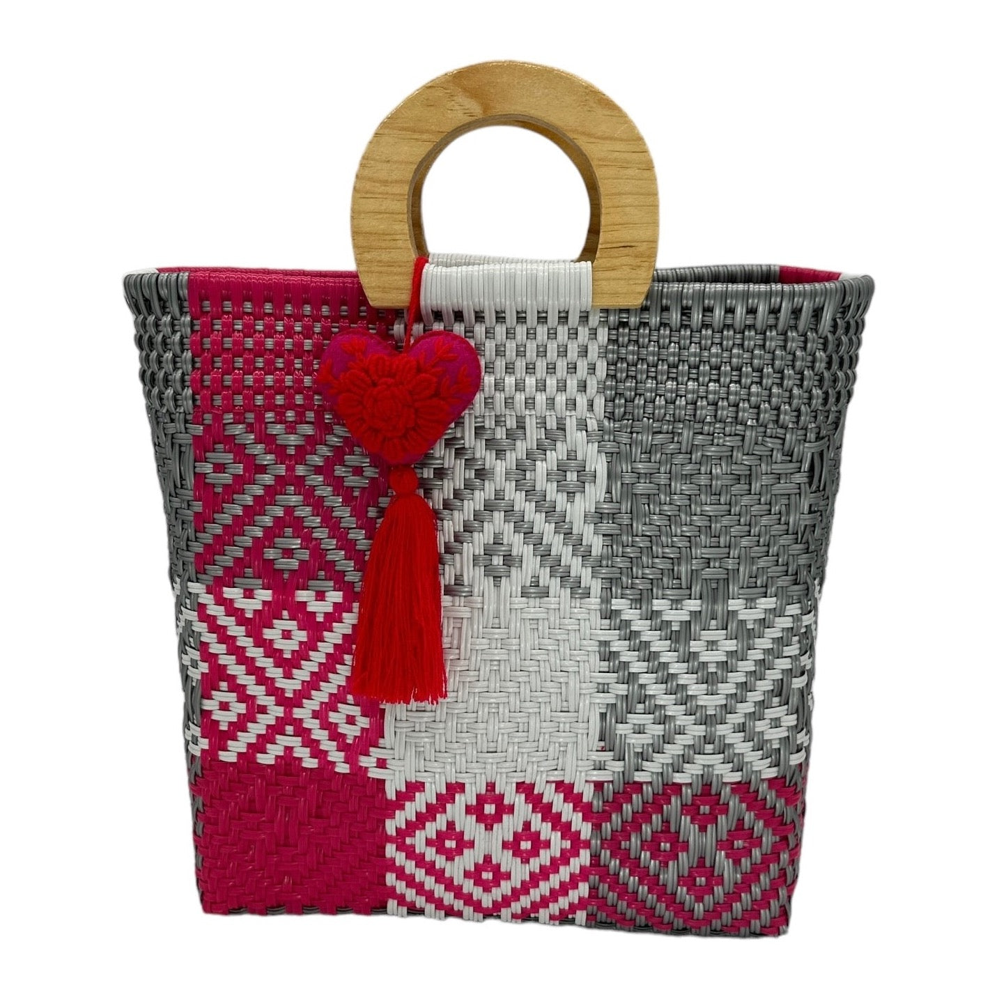 woven tote bag made of recycled plastic with a wooden circular handle. The bag features a patchwork design in pink, gray, and white with geometric patterns. It is adorned with a heart-shaped charm with a tassel.