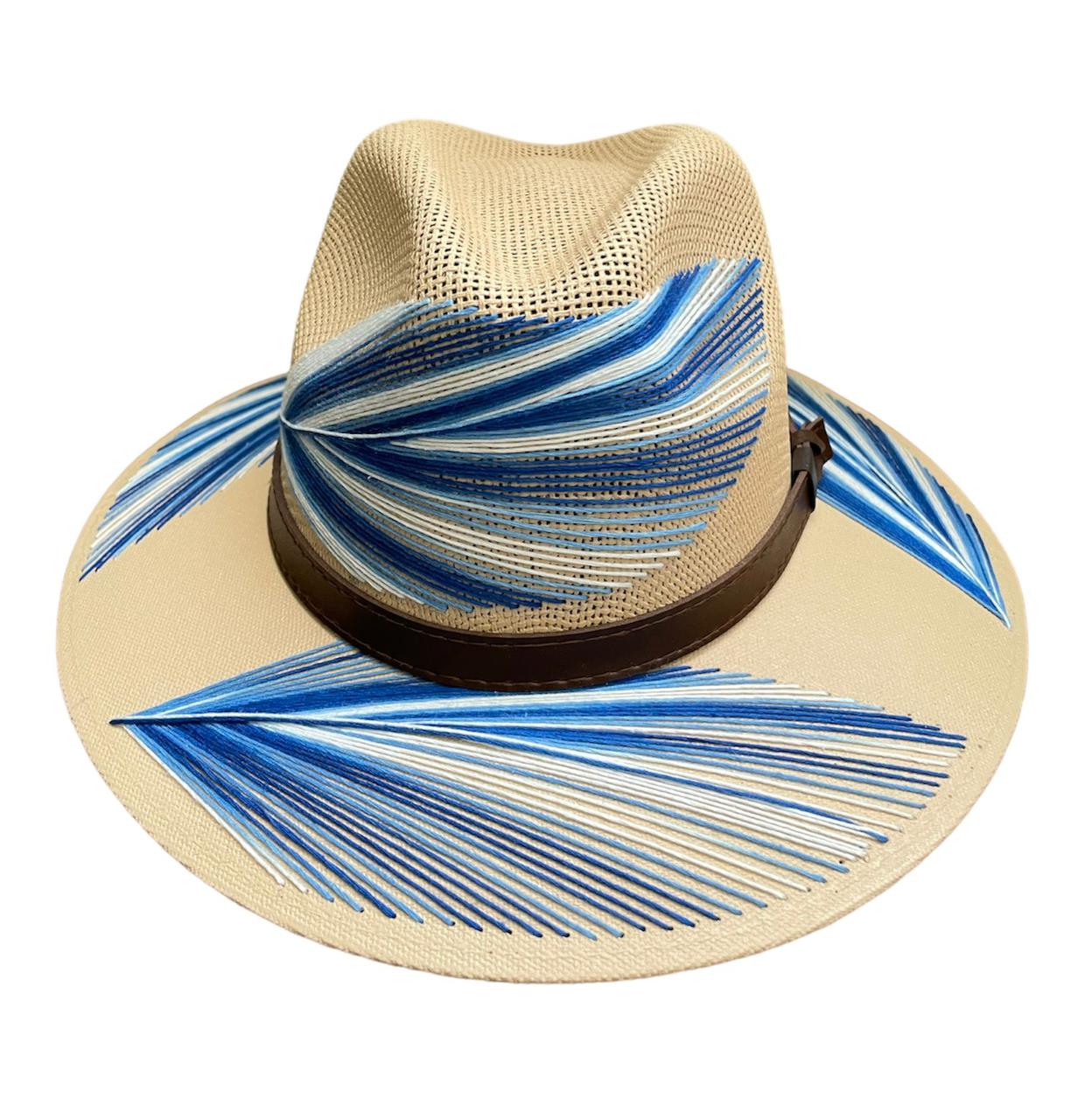 beige hat with a wide brim featuring blue and white painted designs resembling palm fronds. It has a brown leather band around the base of the crown.