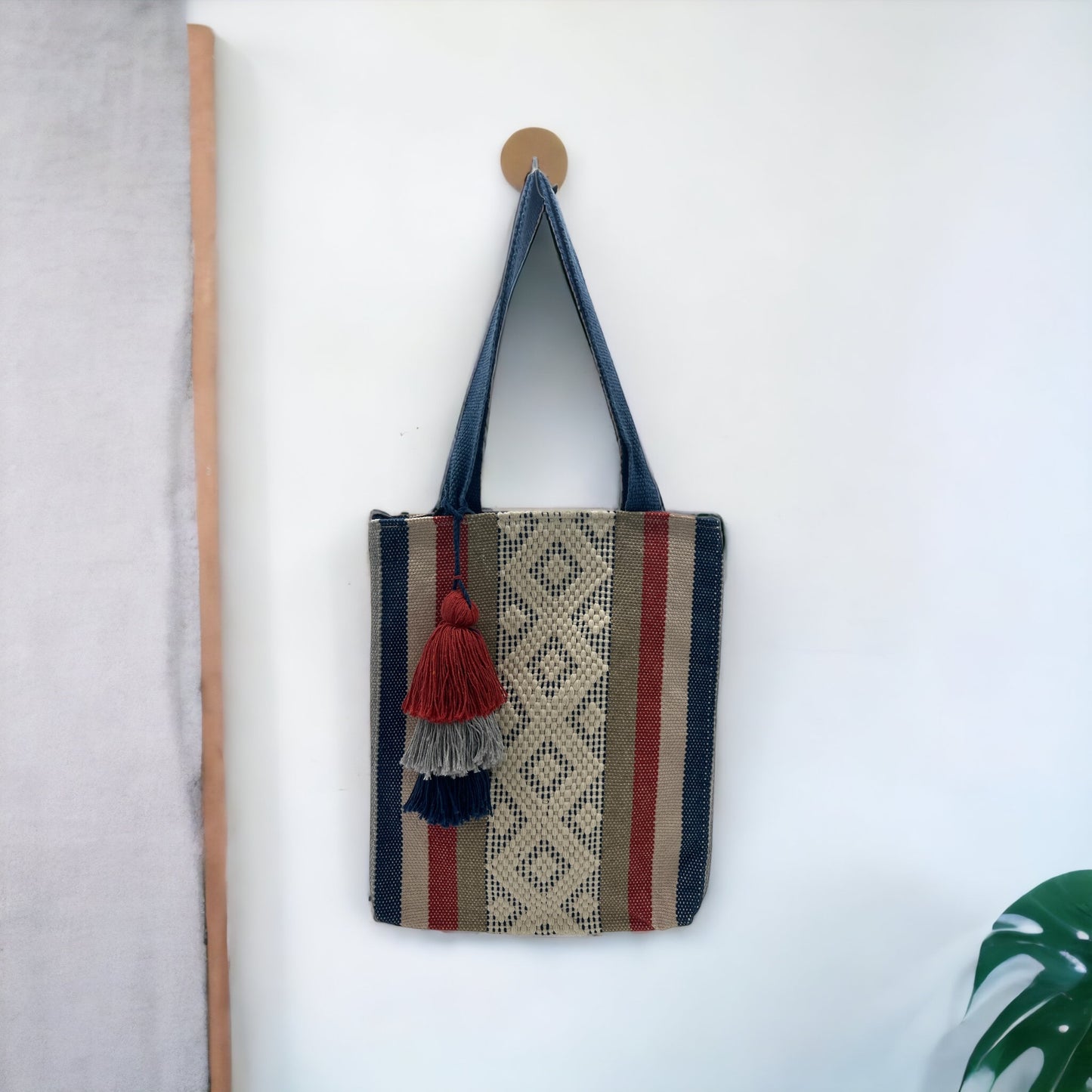 woven tote bag with vertical stripes in blue, beige, and red, featuring a decorative tassel. The bag is hanging on a white wall.