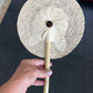 handwoven palm leaf fan with a circular shape, featuring a flower design in the center. The fan is held by a wooden handle.
