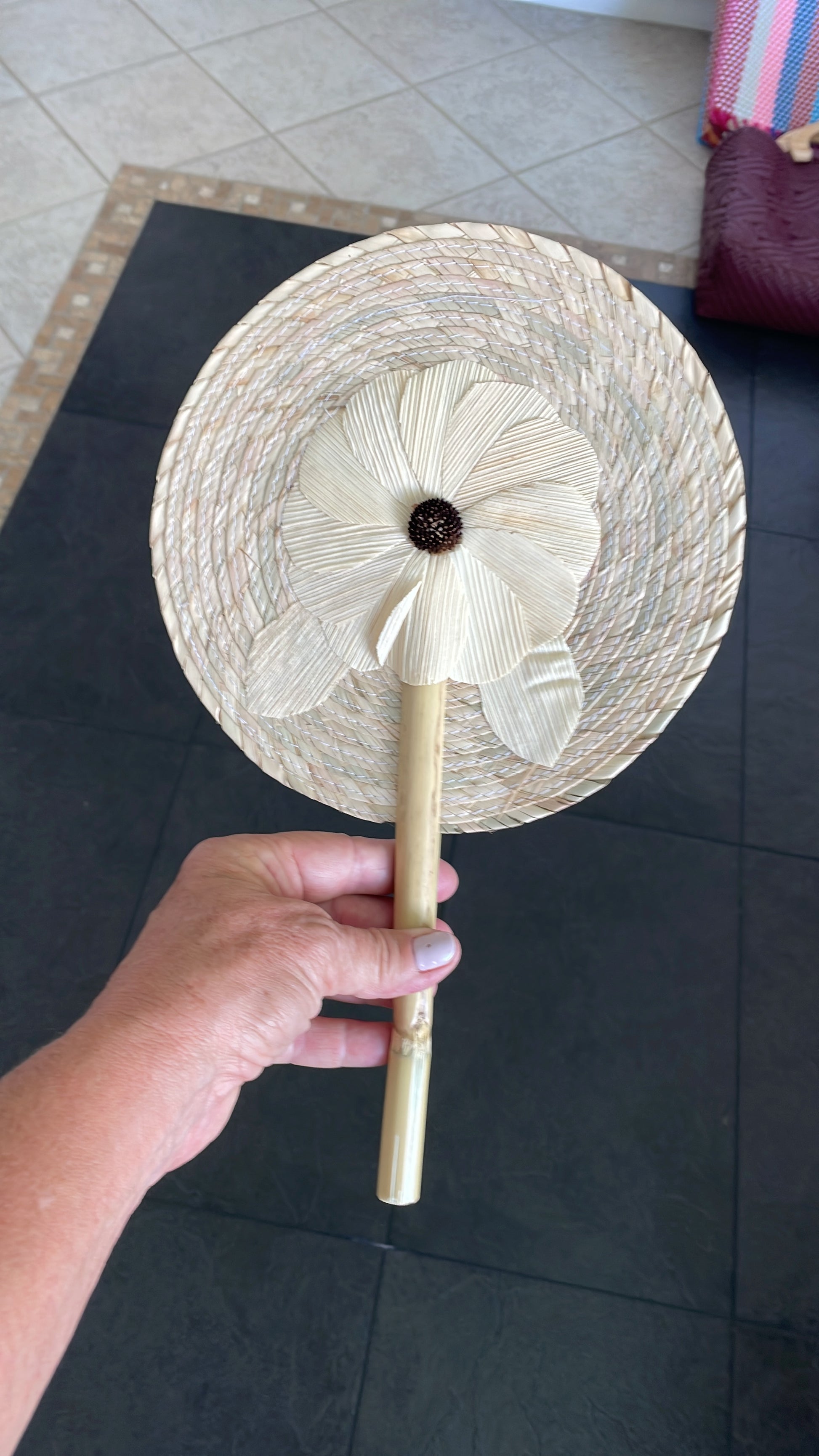 handwoven palm leaf fan with a circular shape, featuring a flower design in the center. The fan is held by a wooden handle.