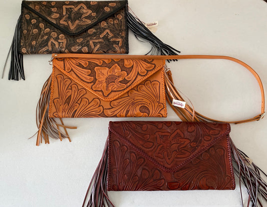 Three leather clutches with floral hand-carved detailing and large side tassels. The clutches are in brown, tan, and burgundy, with snap front closures and lined interiors. One has a long strap for crossbody wear.