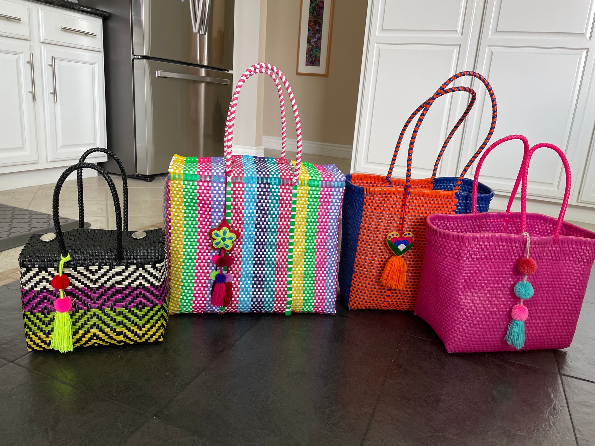 collection of colorful handcrafted bags placed on a kitchen floor. Each bag is made from recycled plastic and features vibrant, multicolored designs. The bags vary in size and color, including patterns such as chevron and checkered, and are embellished with playful decorations like tassels and heart-shaped charms. These bags are ideal for versatile lifestyle activities and make a striking visual statement with their bright hues.