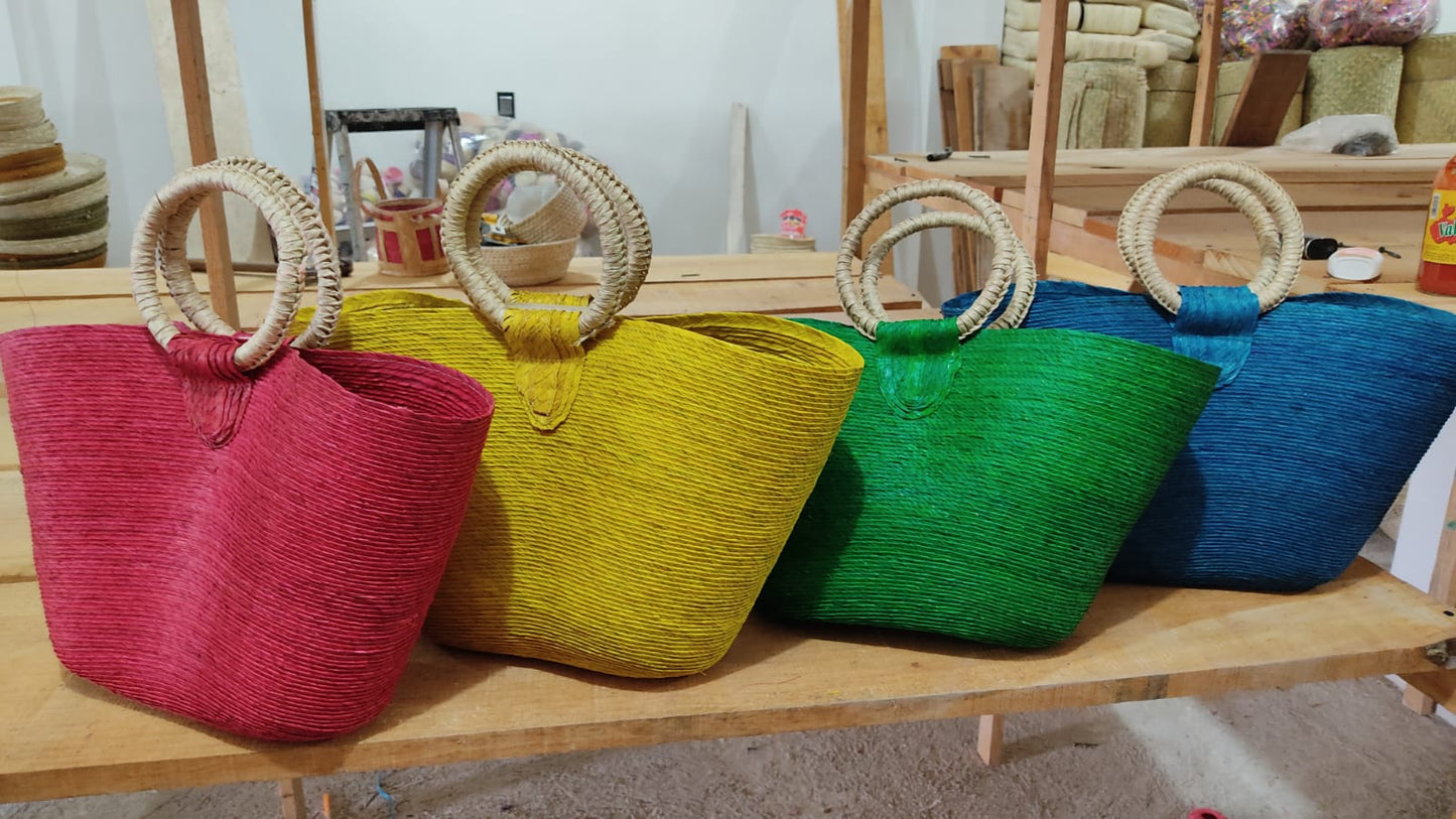 large woven tote bags in red, yellow, green, and blue, each with circular handles. The bags are displayed on wooden shelves in a workshop setting.