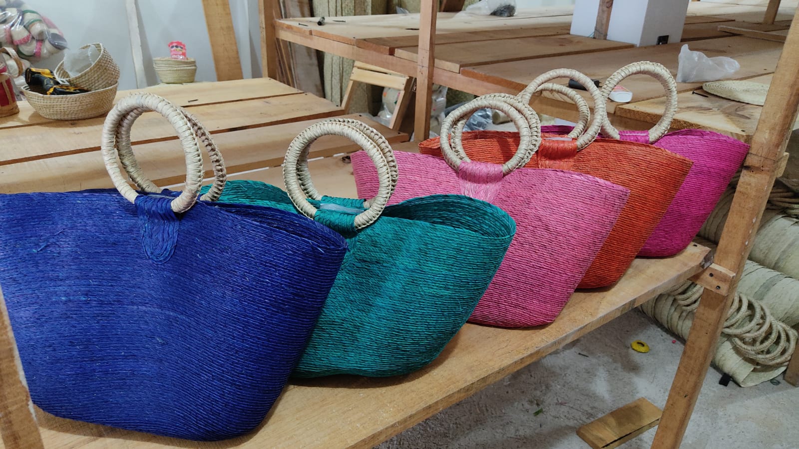 large woven tote bags in various colors (blue, teal, orange, pink, and magenta) with circular handles. The bags are placed on wooden shelves in a workshop setting.