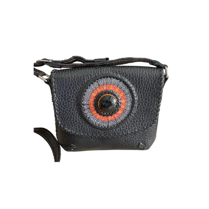 Large black crossbody bag with a circular medallion design on the front. The medallion features intricate patterns in grey, red, and yellow, with a large black center. The bag has a textured leather finish and an adjustable strap.