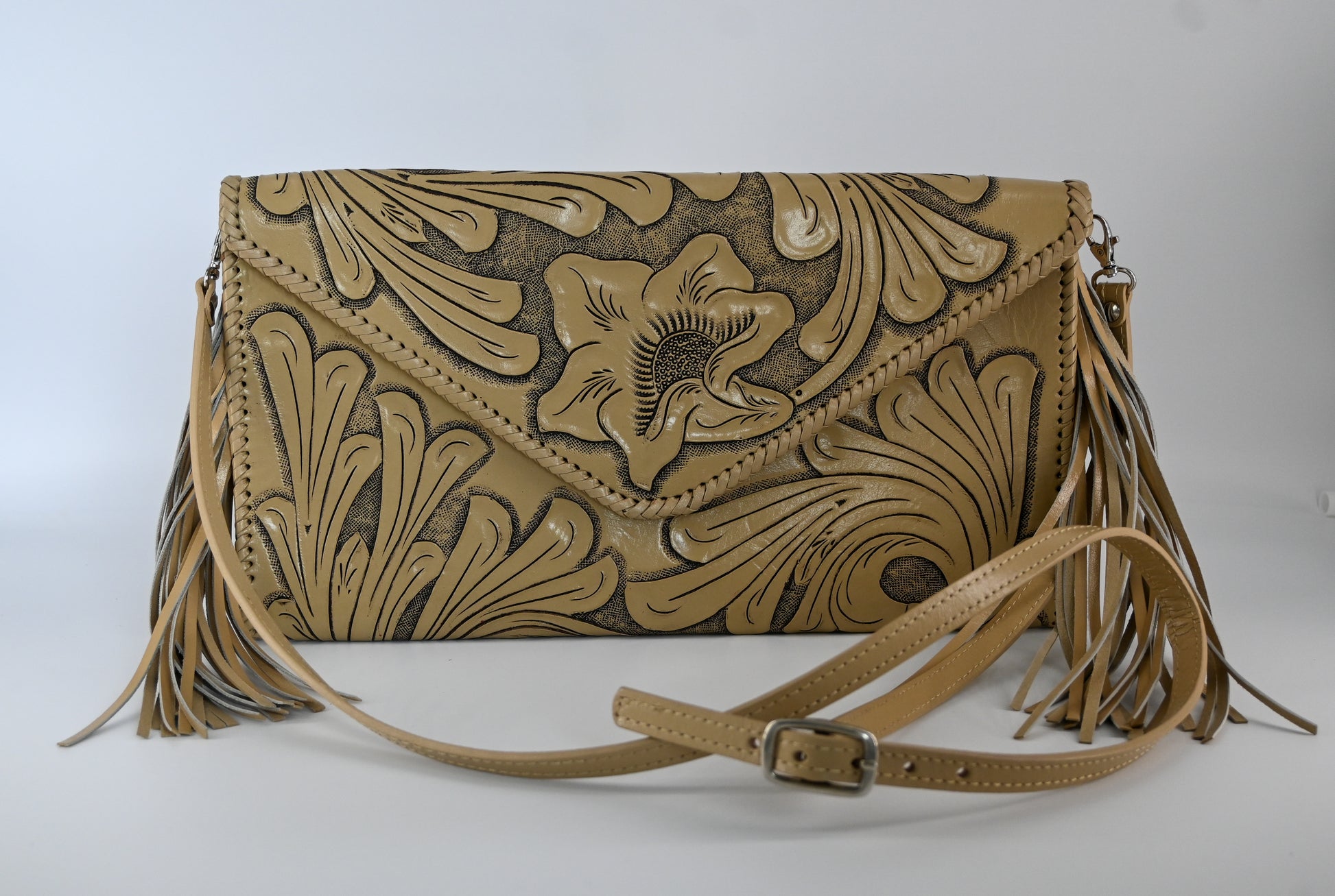 tan leather clutch with intricate floral carvings, long side tassels, and a matching strap. It has a snap front closure.