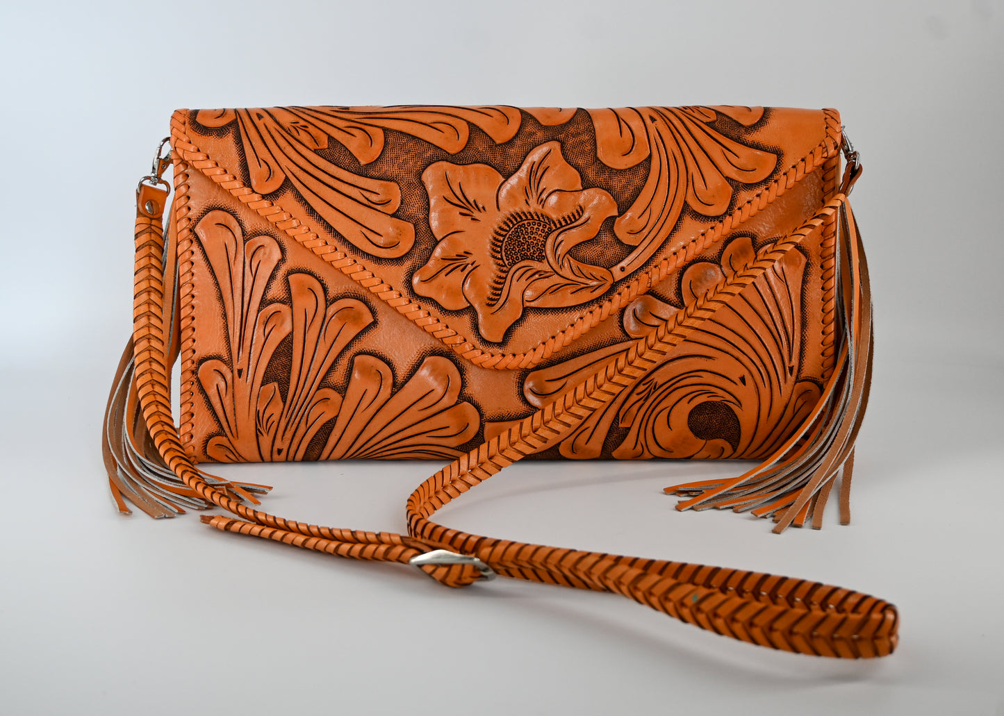 honeyleather clutch with hand-carved floral detailing, large side tassels, and a braided strap. The clutch has a snap