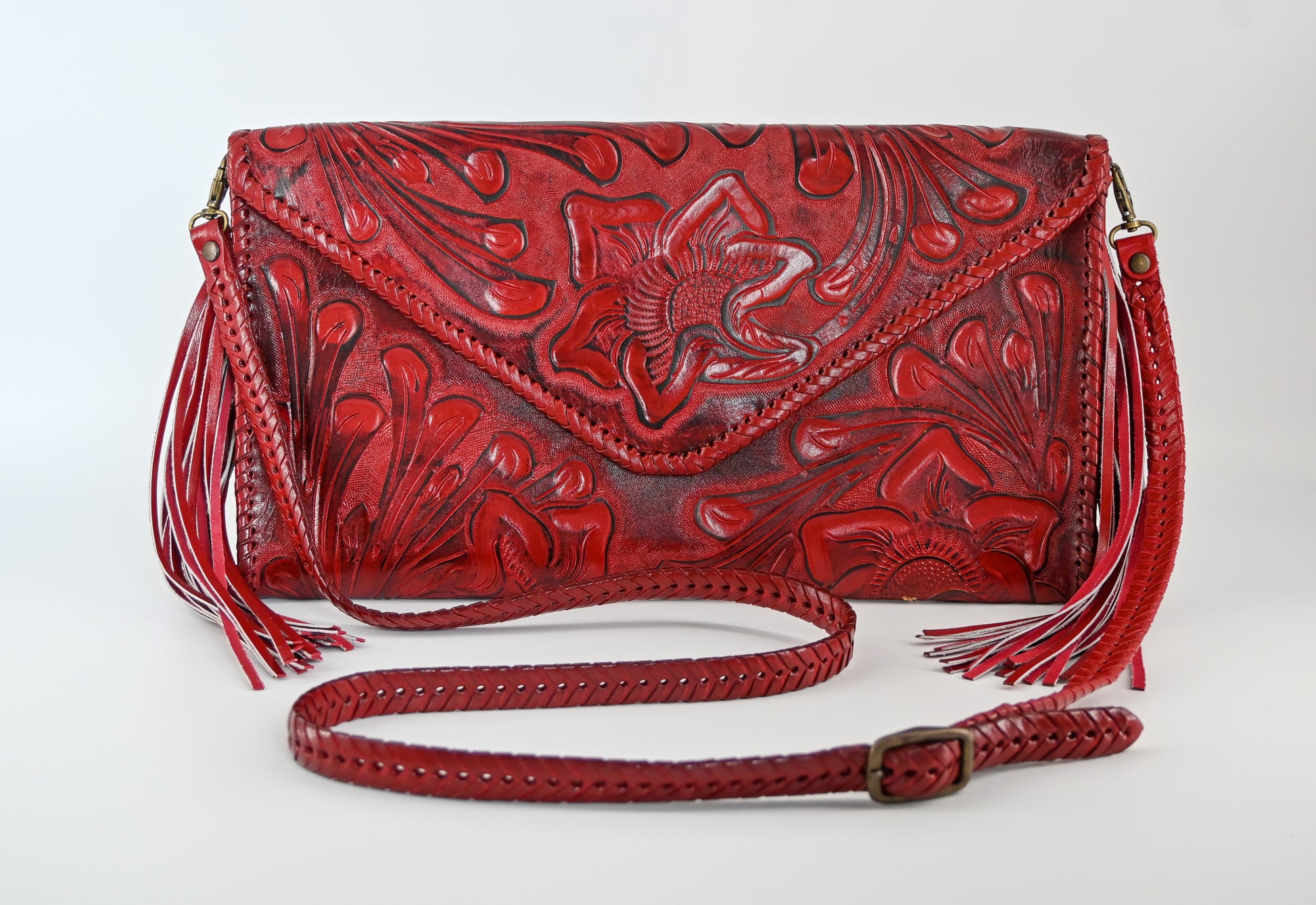 red leather clutch with intricate floral carvings, long side tassels, and a matching braided strap. It has a snap front closure.