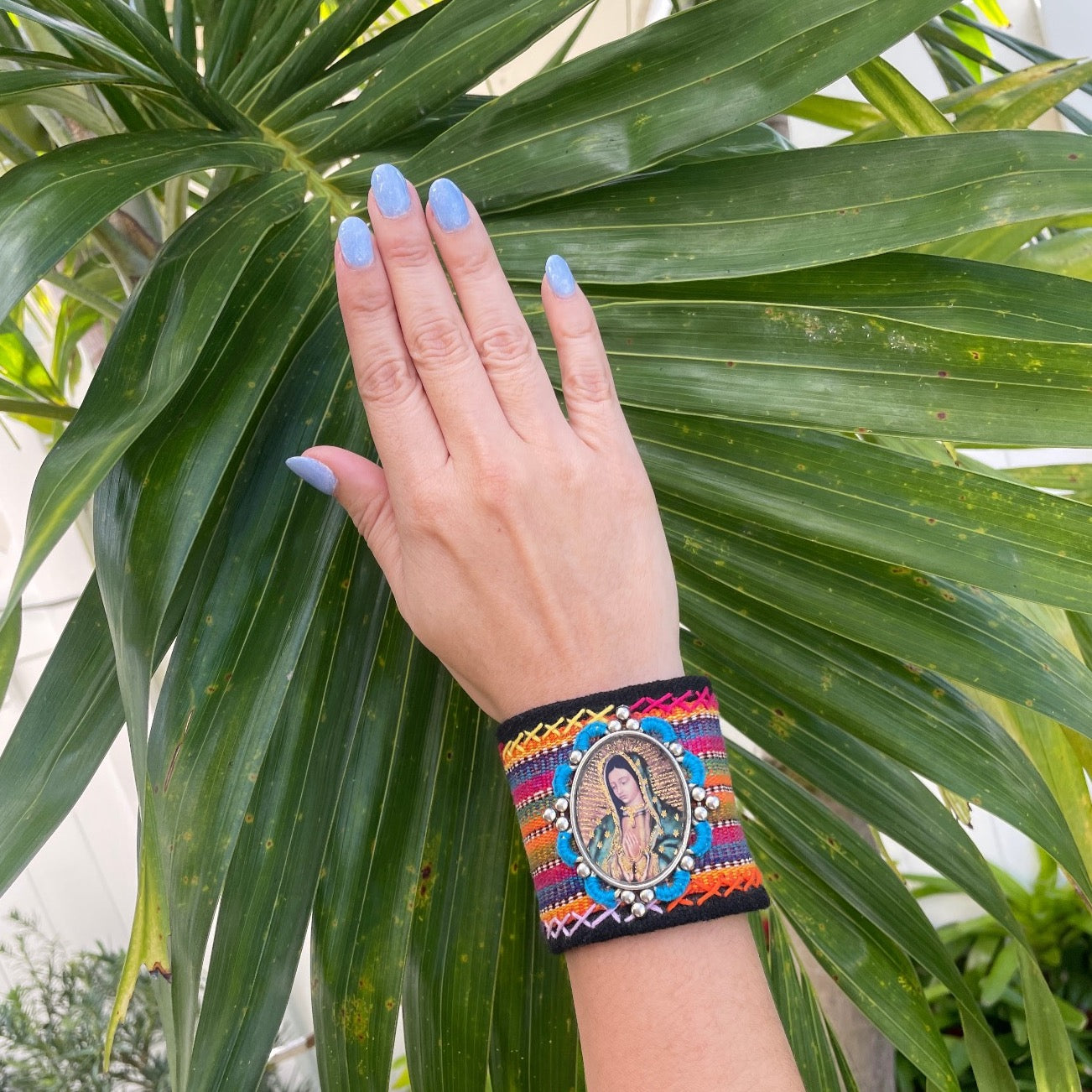 bracelet is decorated with vibrant, textured threads and features a central framed image of The Virgin Mary. It is secured with two magnetic snaps.