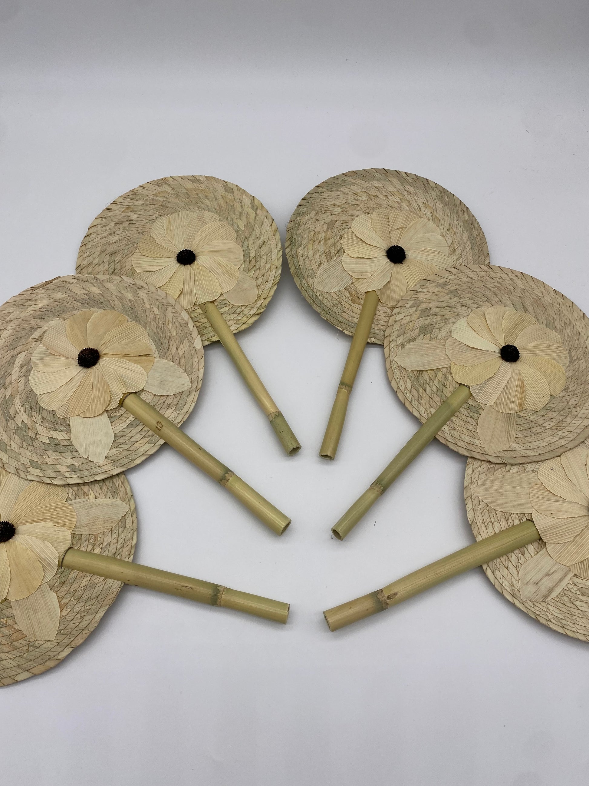 handwoven palm leaf fan with a circular shape, featuring a flower design in the center. The fan is held by a wooden handle.