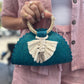 small, handwoven, teal-colored crescent handbag made of palm leaves, featuring a braided white tassel and a woven handle.