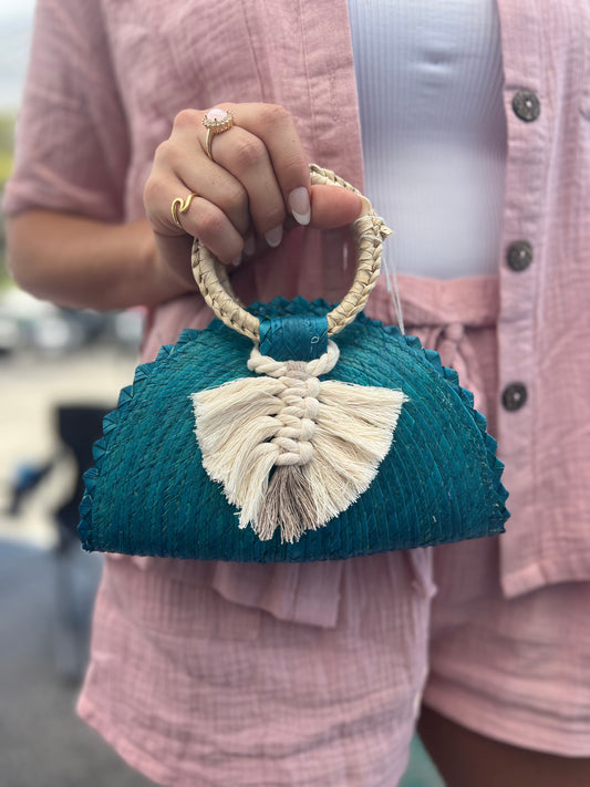 small, handwoven, teal-colored crescent handbag made of palm leaves, featuring a braided white tassel and a woven handle.