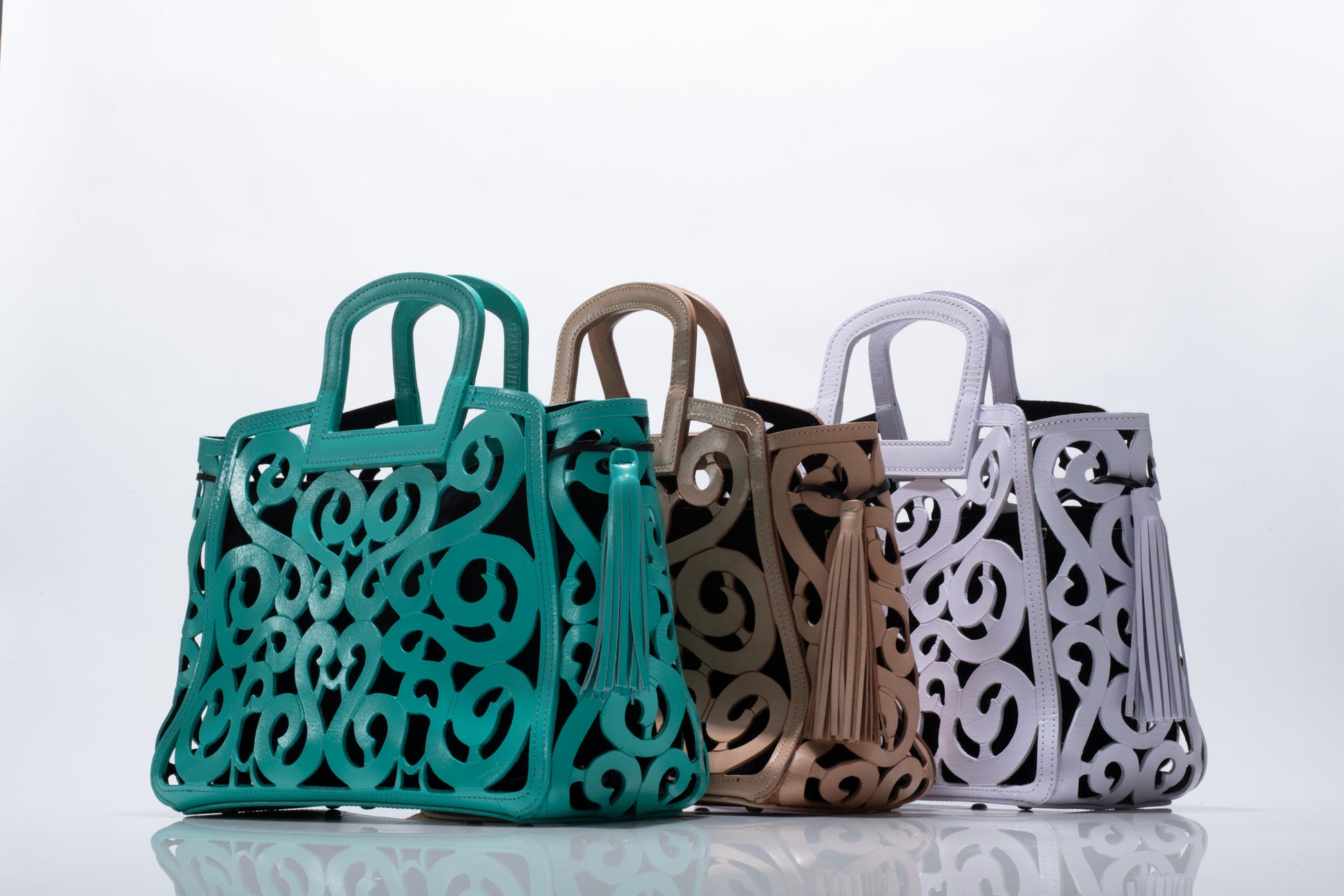 Three leather tote bags with intricate cut-out designs and tassel accents are displayed side by side. The bags are in turquoise, nude, and white colors.