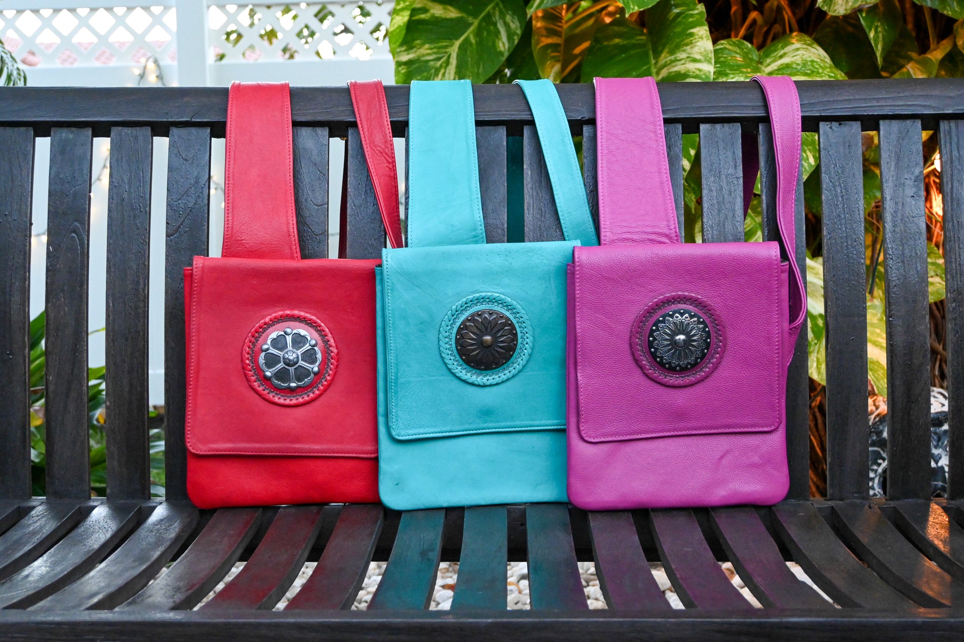 Three colorful leather crossbody bags with wide straps and circular decorative medallions on the front flap, displayed on a wooden bench. The bags are red, turquoise, and purple.