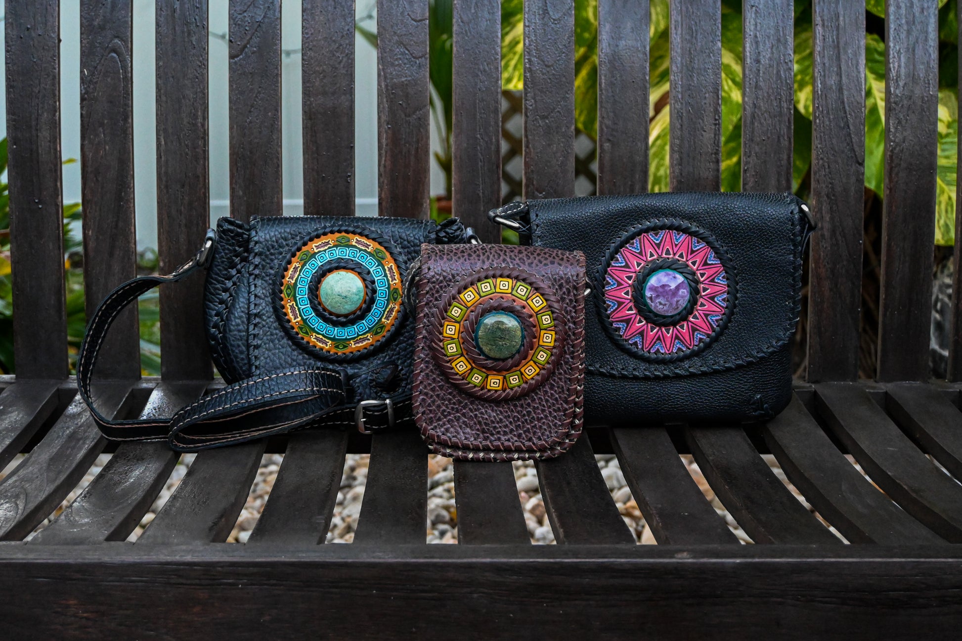 Three small,medium and large crossbody bags with circular medallions on the front are displayed on a wooden bench. The bags are black and brown, each featuring unique colorful geometric designs on the medallions. The bags have adjustable straps and detailed stitching.