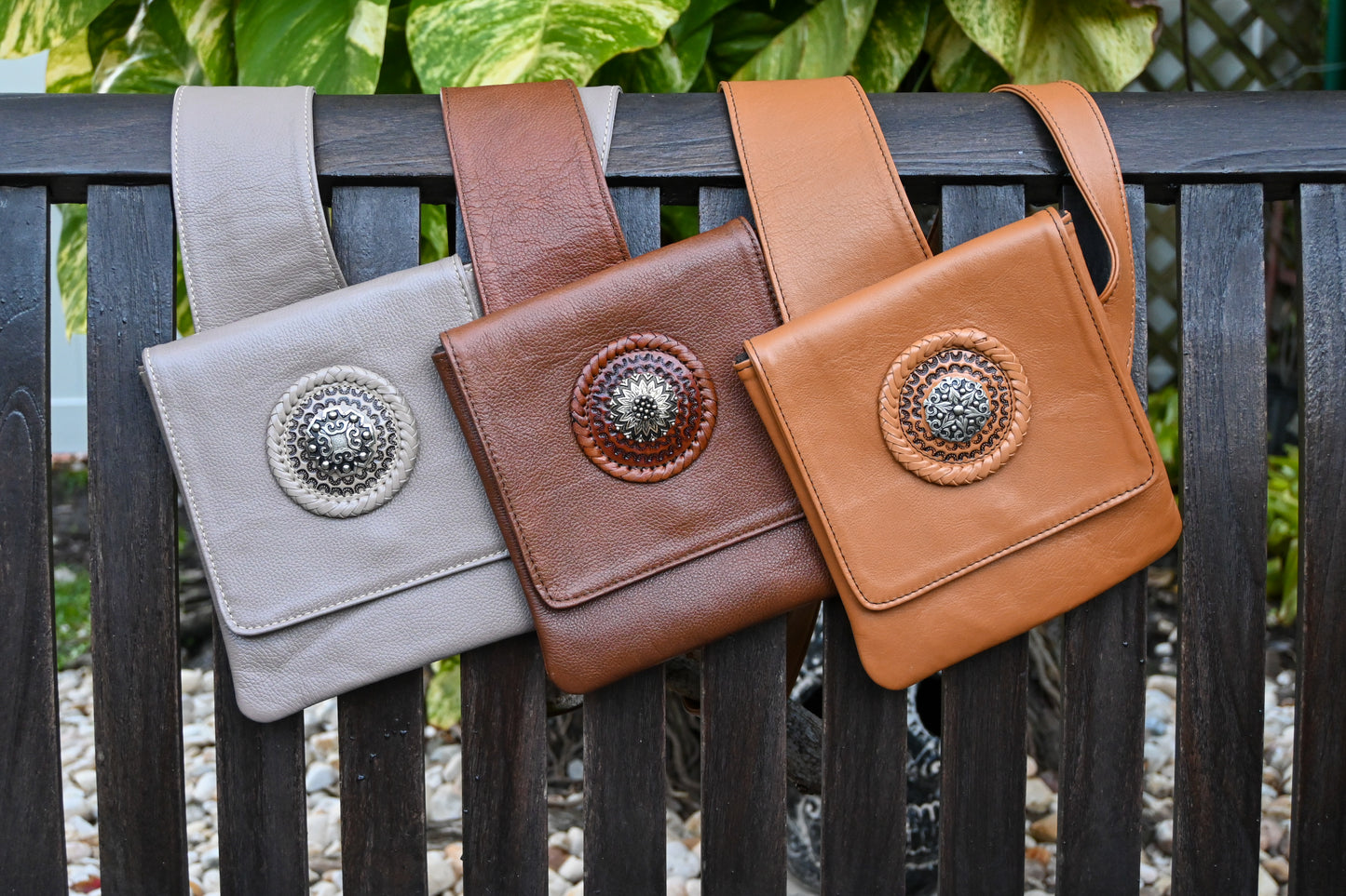 Three leather crossbody bags with wide straps, each featuring a circular decorative medallion on the front flap. The bags are in beige, brown, and tan colors, and are displayed hanging on a wooden bench.