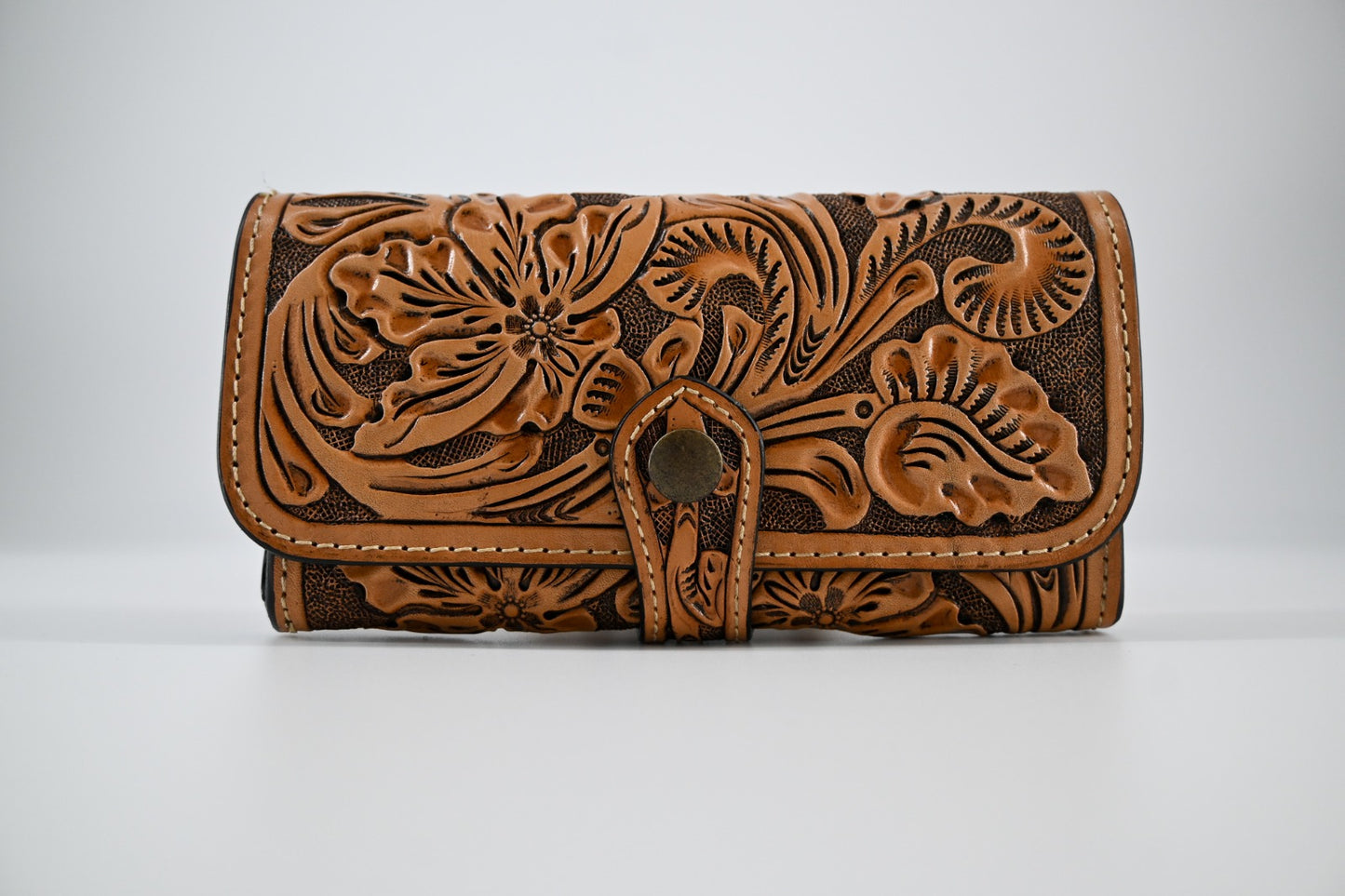 tan leather wallet with an intricate hand-carved floral design on the front. It features a button closure and detailed stitching along the edges.