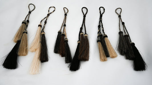 set of hand-braided horse hair tassels in various shades of black, brown, and blonde, each featuring a loop for attachment. The tassels are arranged in a row against a plain background.