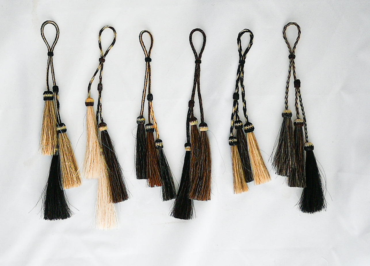 set of hand-braided horse hair tassels in various shades of black, brown, and blonde, each featuring a loop for attachment. The tassels are arranged in a row against a plain background.