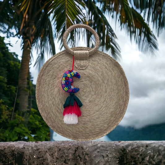 round woven handbag made of natural fibers with a circular handle. The bag is adorned with a colorful crescent moon charm with floral patterns and tassels 